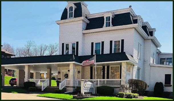 Large white Victorian-style house with a covered porch, dark shutters, and manicured lawn under a blue sky