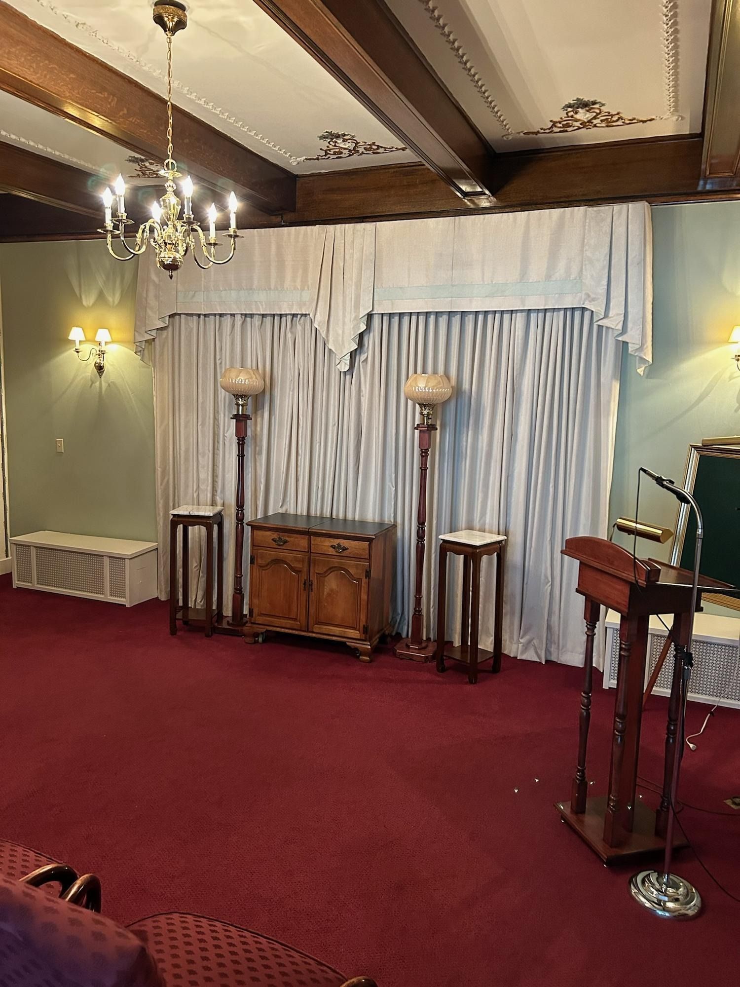Formal room with red carpet, chandelier, curtained backdrop, wooden cabinet, and podiums on a stage.