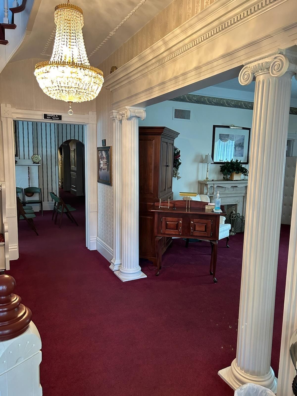 Elegant hallway with ornate columns, crystal chandelier, burgundy carpet, and a wooden desk in the background