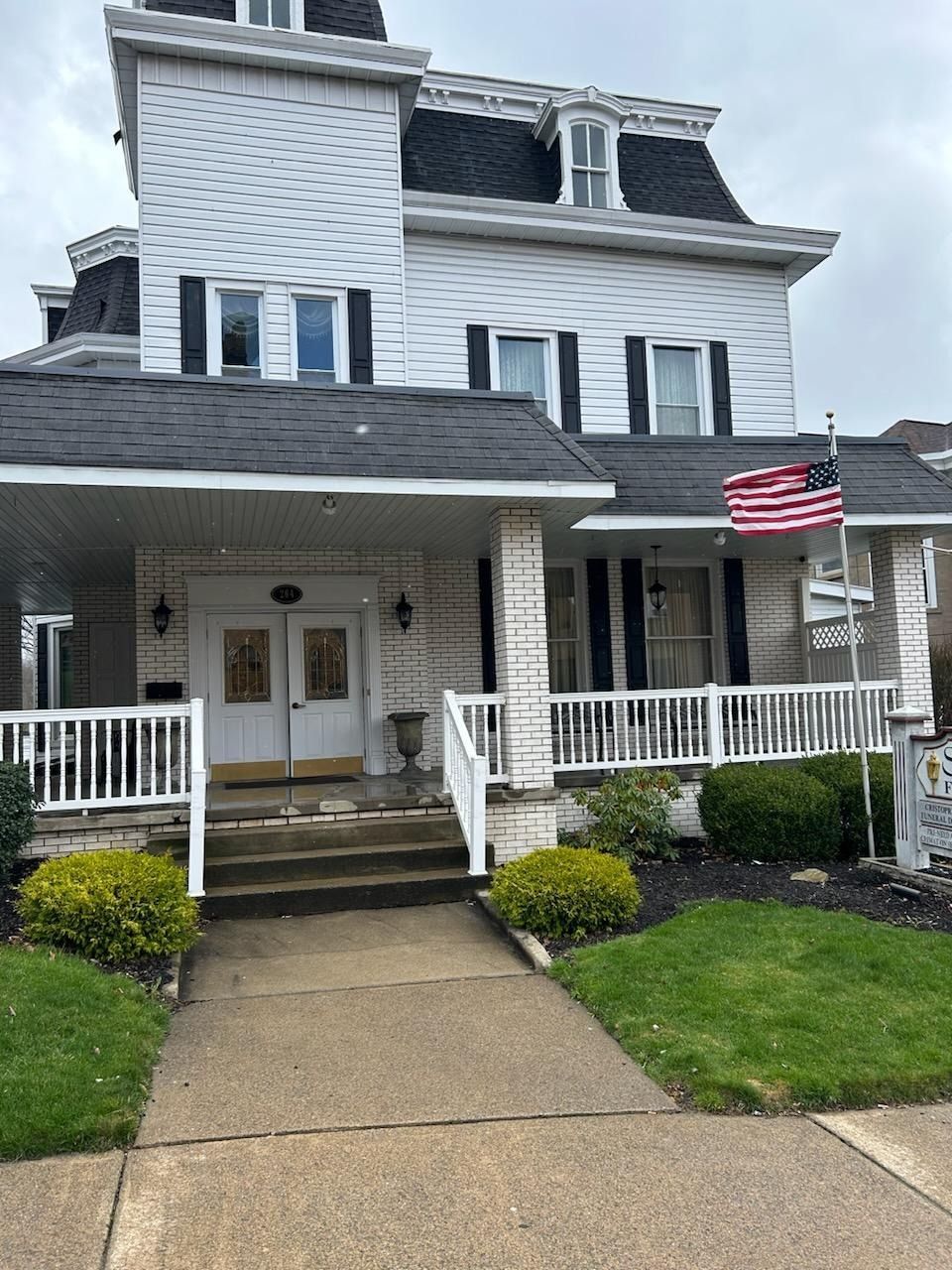 White house with a covered porch, blue shutters, American flag, and a walkway leading to the front door.
