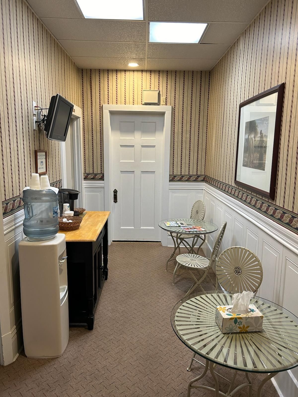 Narrow waiting room with white door, patterned wallpaper, glass tables, chairs, and water cooler
