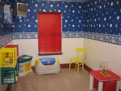 Child's playroom with blue patterned wallpaper, red blinds, toys, and a yellow chair.