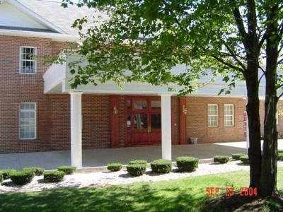 Brick building with white columns and red doors under a canopy; small bushes line the entrance; tree in the right foreground.