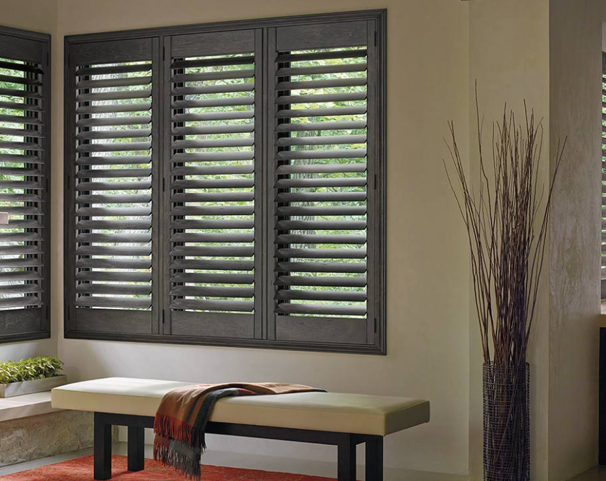 Dark shutters cover a window above a bench with a blanket, and a vase with branches.