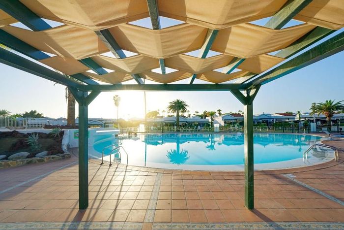 A shaded pergola overlooks a bright blue swimming pool on a sunny day at a resort.