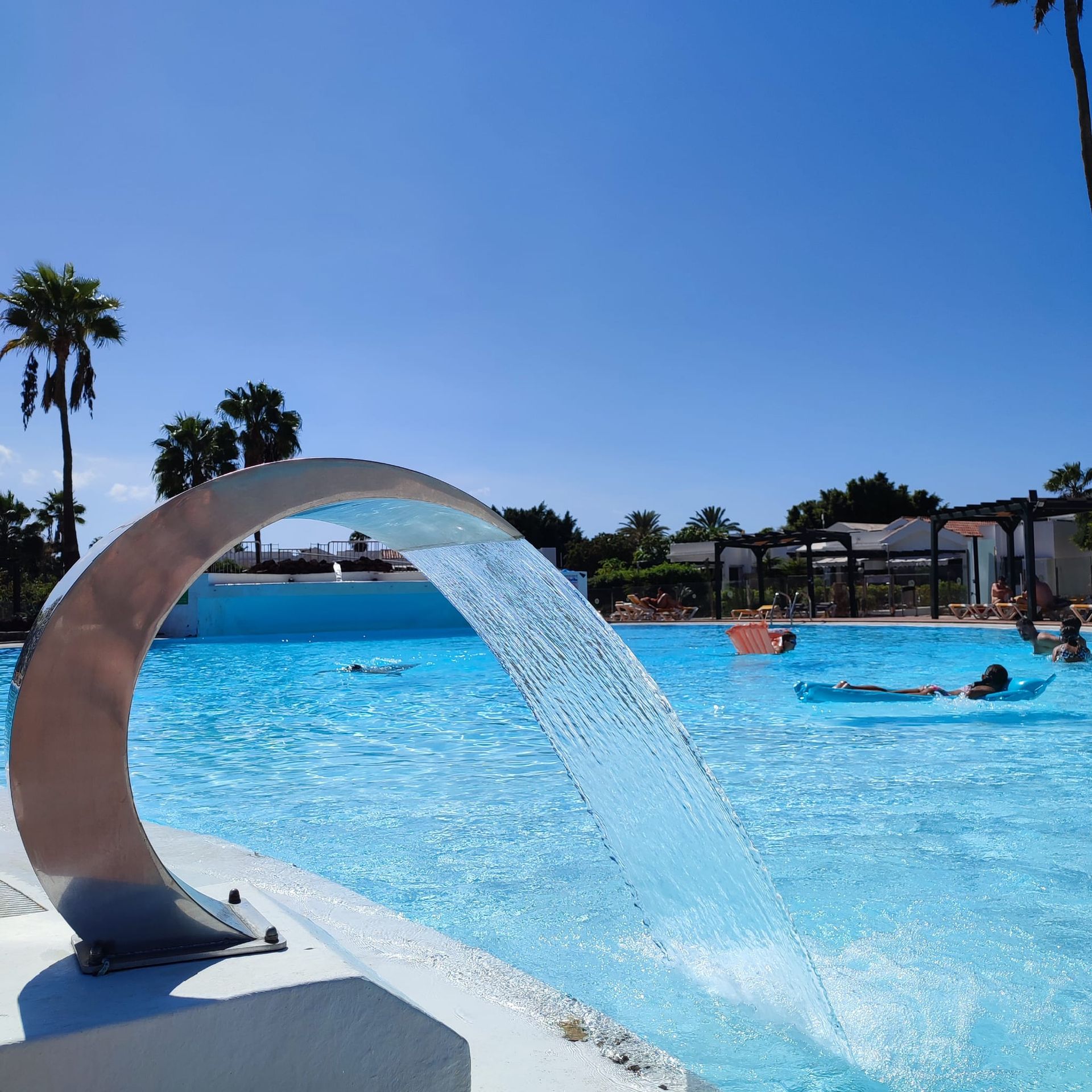A curved metal water feature pouring water into a bright blue sunny swimming pool with people floating in the distance.