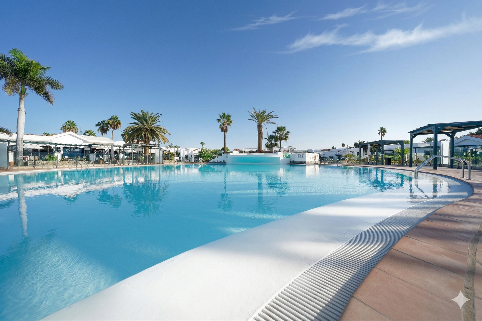 A bright, expansive swimming pool under a clear blue sky, surrounded by palm trees and resort structures.