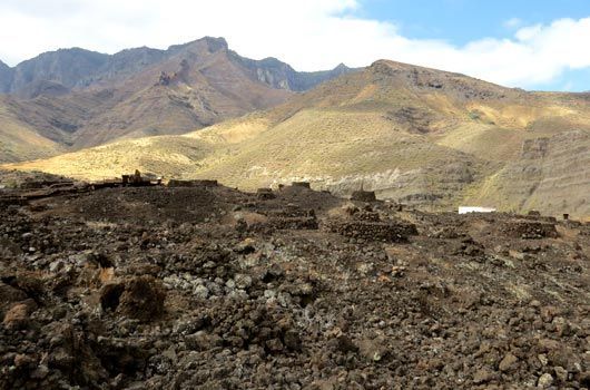 A rocky, arid volcanic landscape in the foreground, with large, rugged mountains rising against a blue, cloudy sky.