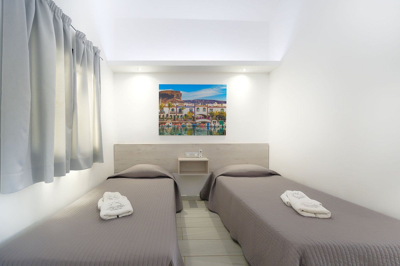 A small hotel bedroom with two single beds featuring gray covers, white linens, and a colorful landscape painting.