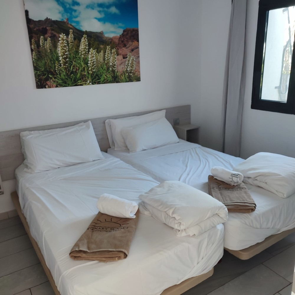 Two twin beds with white linens, folded towels, and a framed nature print on a white wall in a simple bedroom.