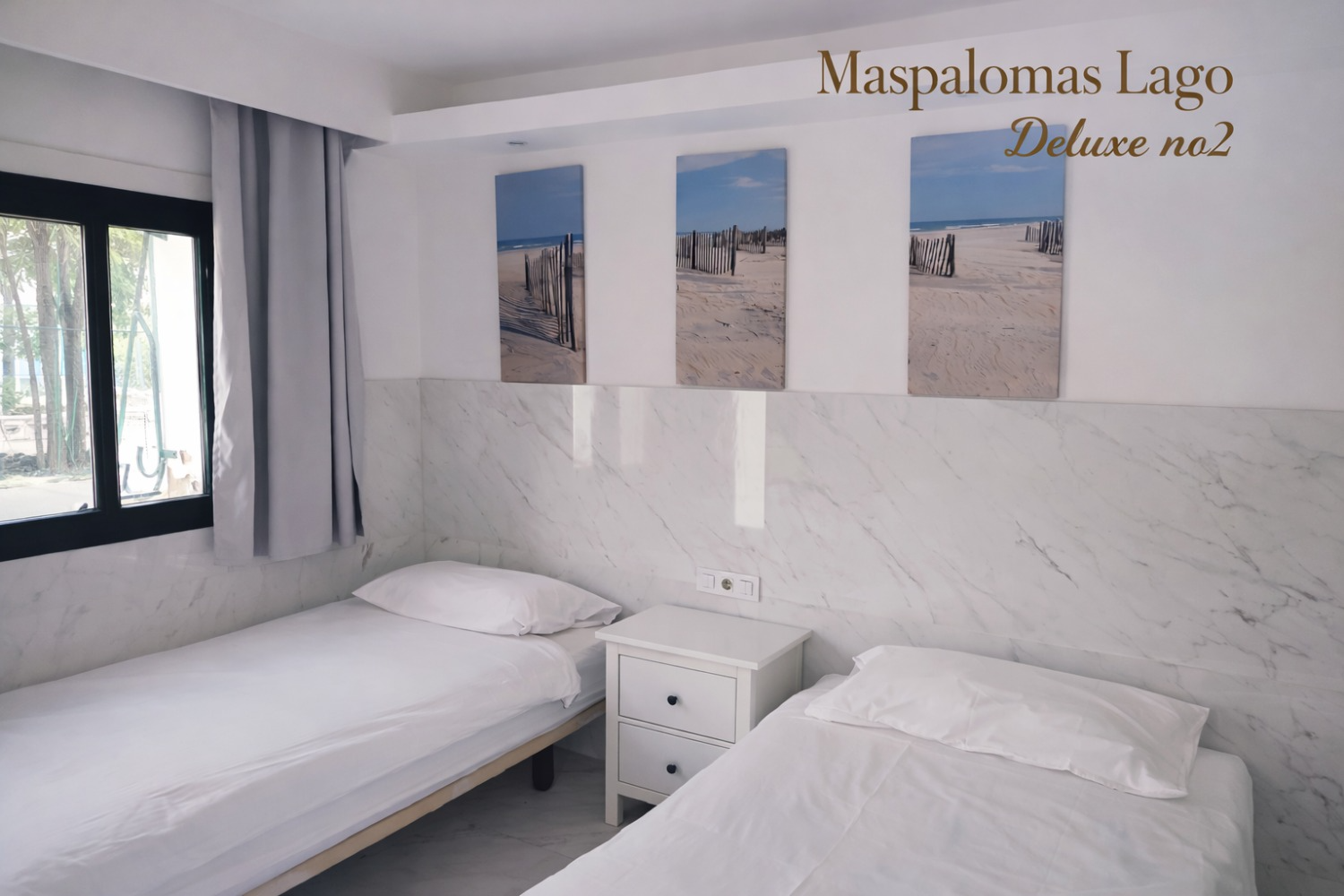 A hotel bedroom with two twin beds separated by a white nightstand, marble-style walls, and three beach artwork panels.