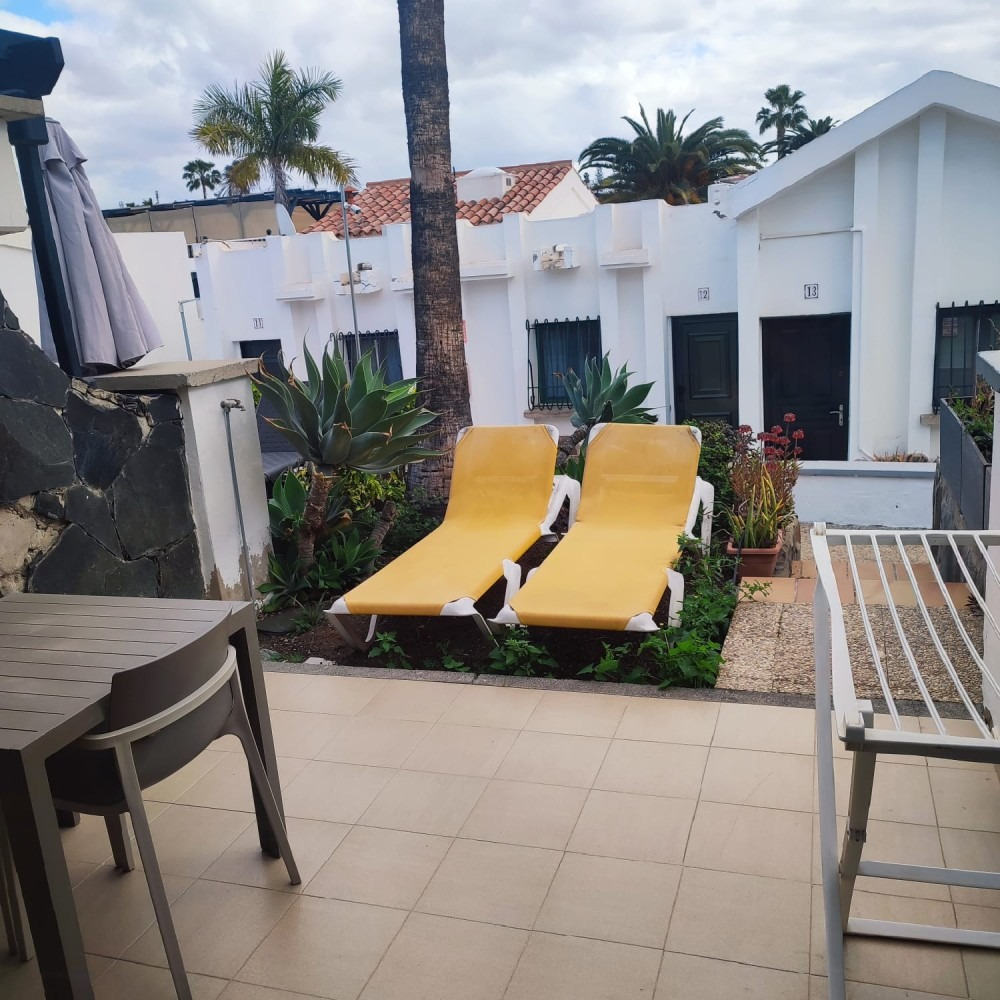 Two yellow lounge chairs sit on a patio area next to a small garden, with white vacation rental units in the background.
