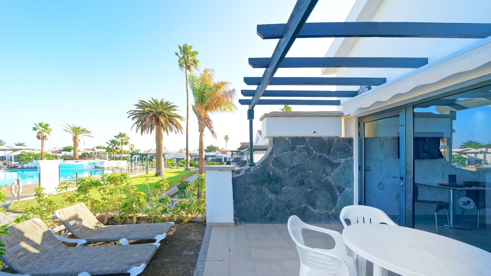 A sunny patio with lounge chairs, a table, and chairs, overlooking a resort pool and palm trees under a clear blue sky.