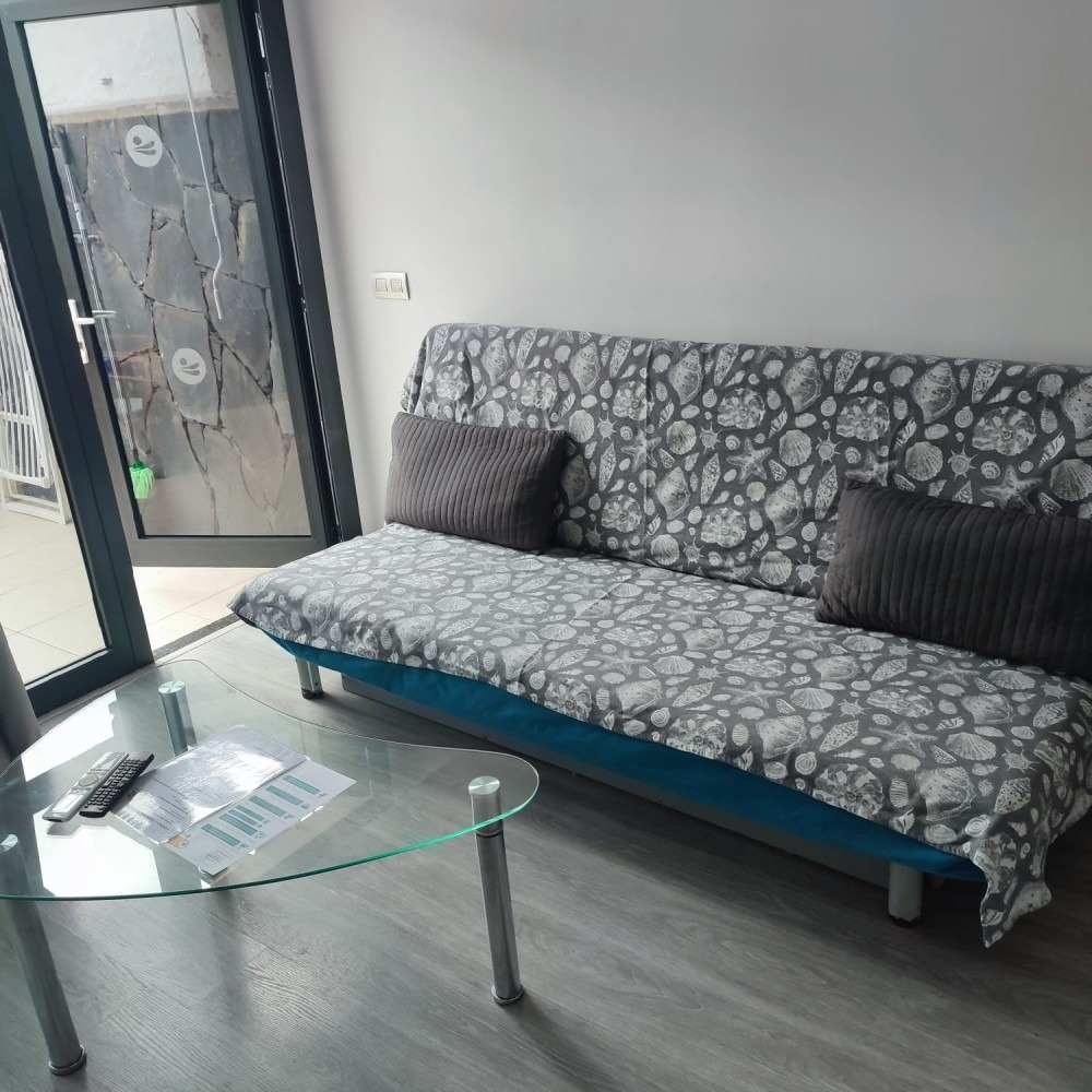A sofa with a gray leaf-print cover and two dark cushions sits beside a glass coffee table in a room with a patio door.
