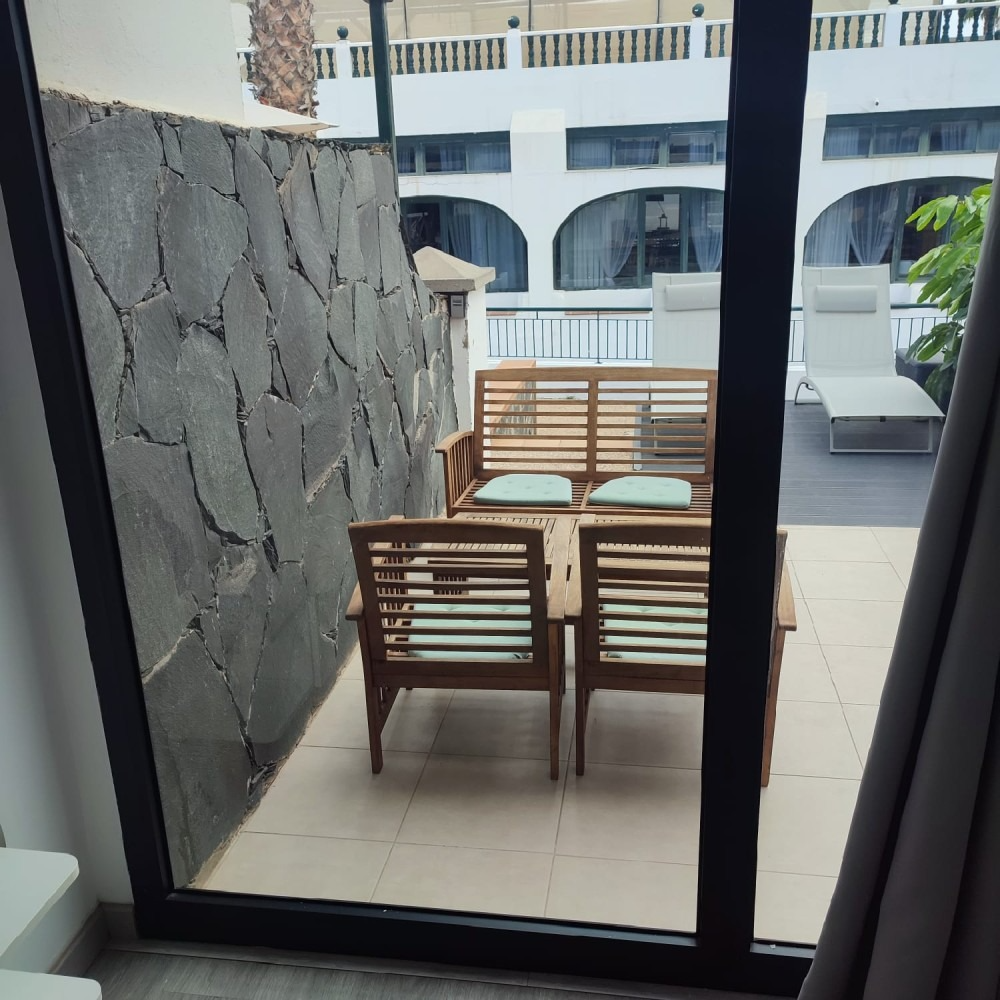 A patio viewed through a glass door, featuring wooden furniture, light cushions, and a stone wall against a hotel complex.