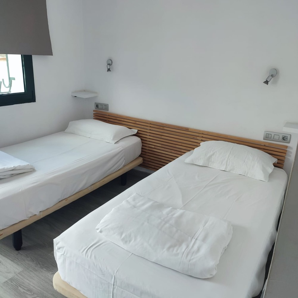 Two twin beds with white linens in a minimalist bedroom with a slatted wooden headboard and white walls.