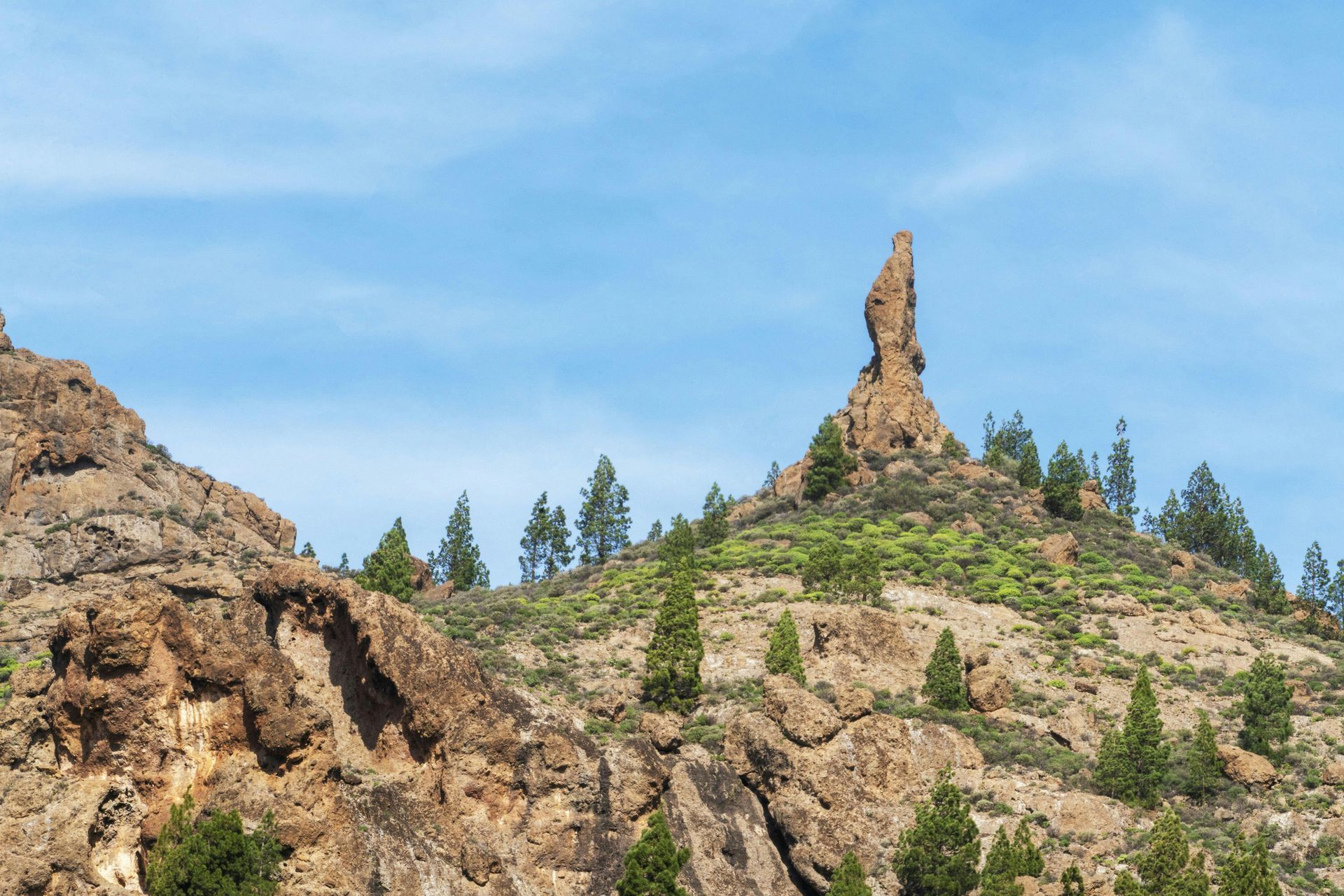 A tall, thin rock formation rises prominently from a sparse, pine-covered hillside against a clear blue sky.