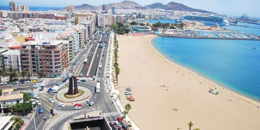 Aerial view of Las Canteras beach in Las Palmas, featuring a roundabout, city buildings, and a cruise ship in the harbor.