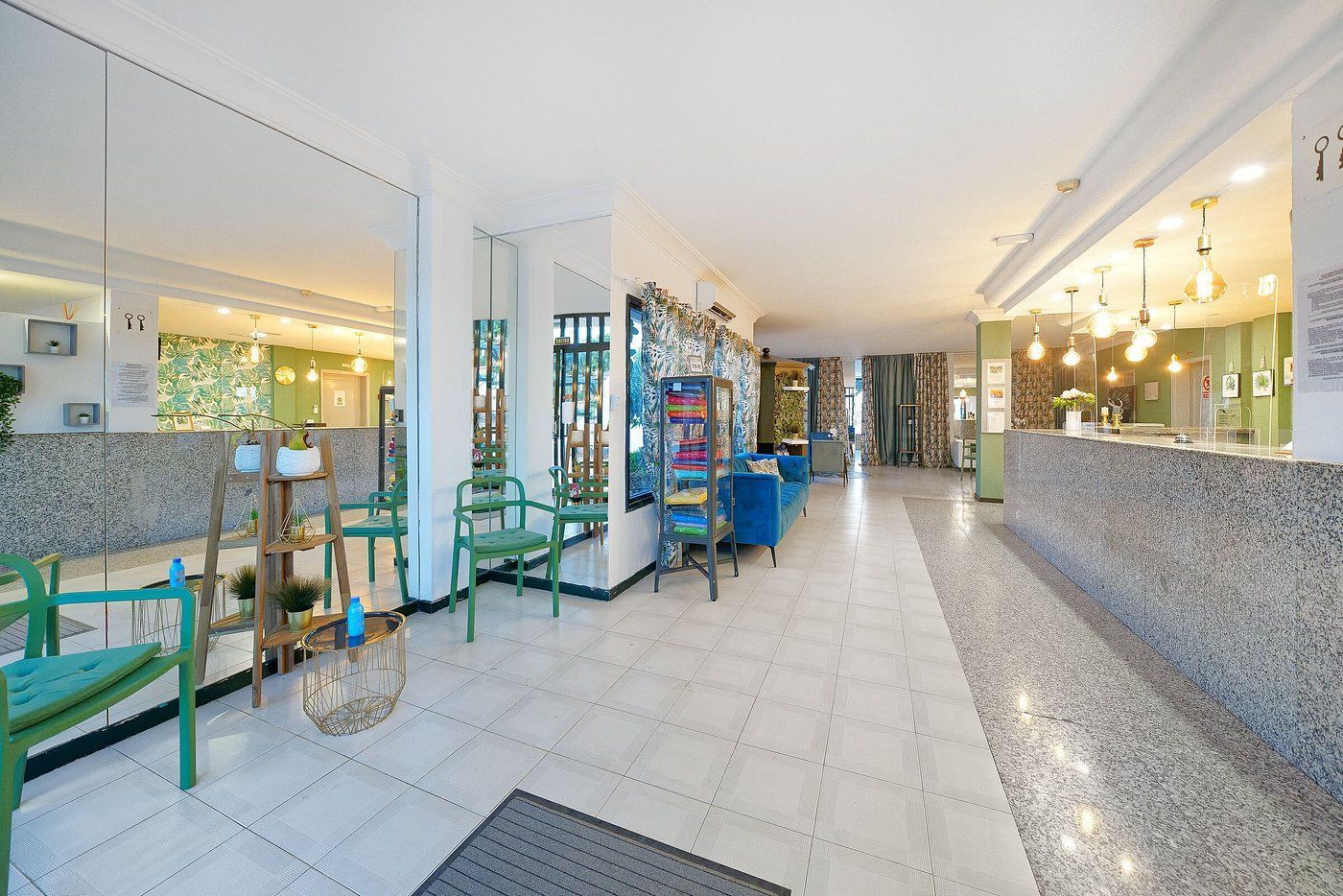 A brightly lit hotel lobby with tile floors, a stone-front reception desk, blue furniture, and mirrored walls.