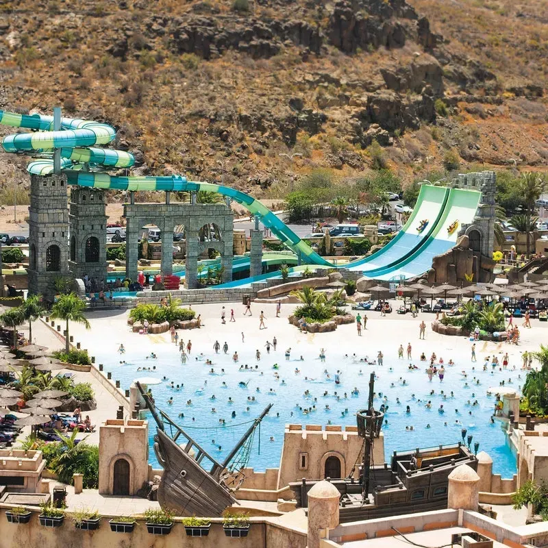 A crowded water park with a turquoise wave pool, a large pirate ship play structure, and high, winding green water slides.