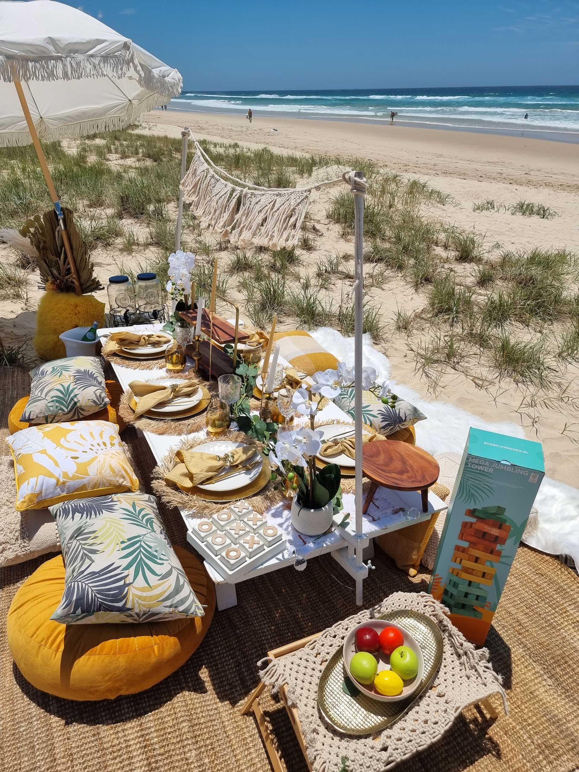 Beach picnic setup with low table, pillows, food, and umbrella on a sunny day.