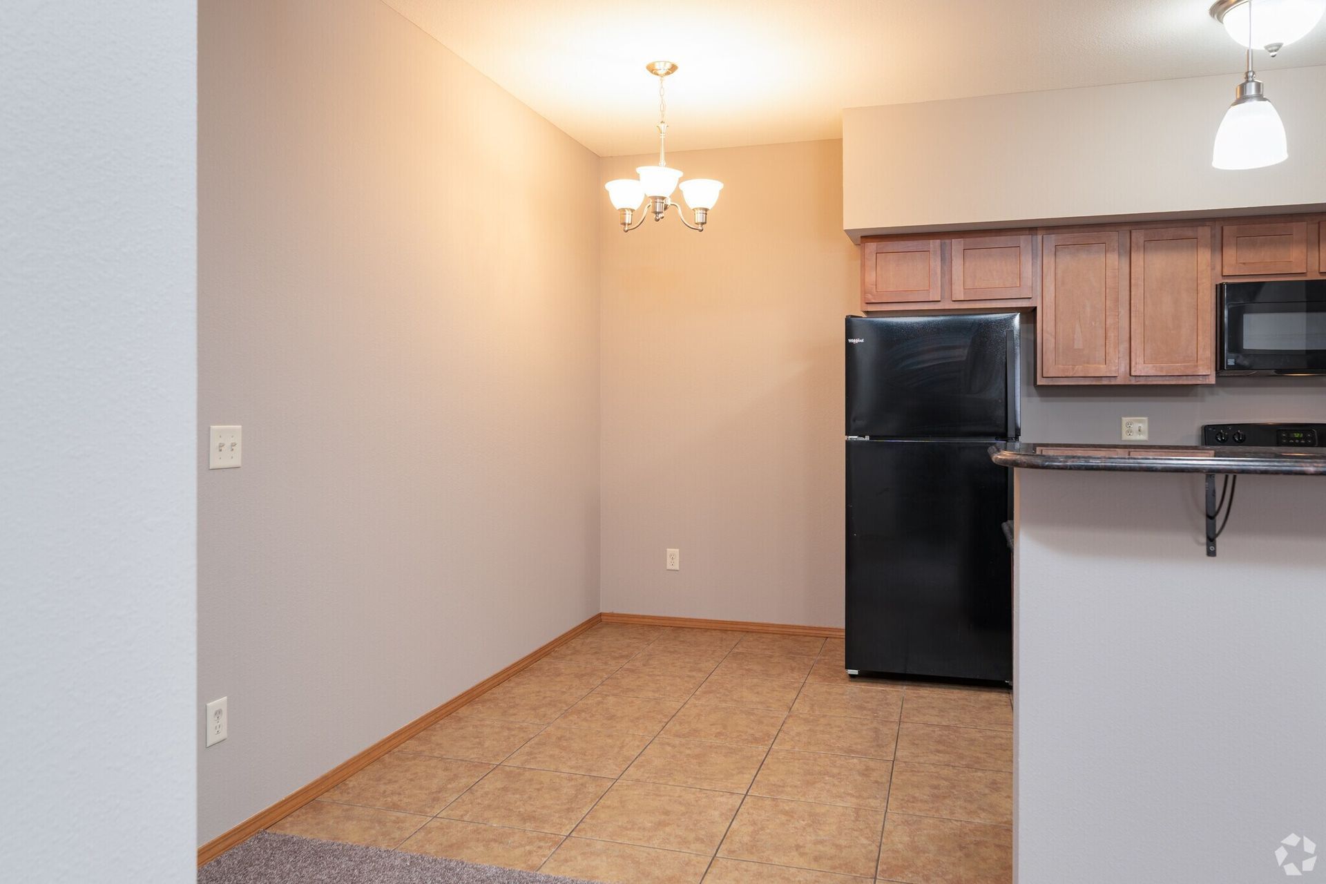 Dining area with black refrigerator, light brown cabinets, chandelier, and beige walls.