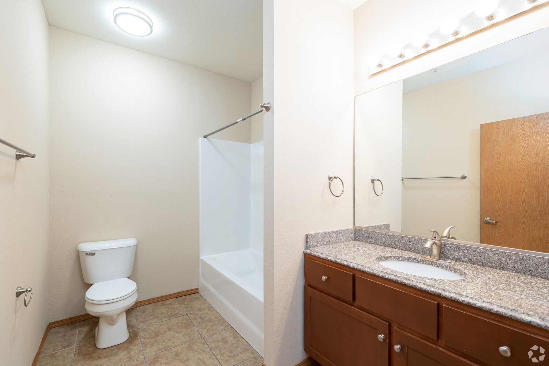 Bathroom with toilet, tub/shower, vanity, and large mirror. Neutral tones, brown cabinetry, and tile floor.