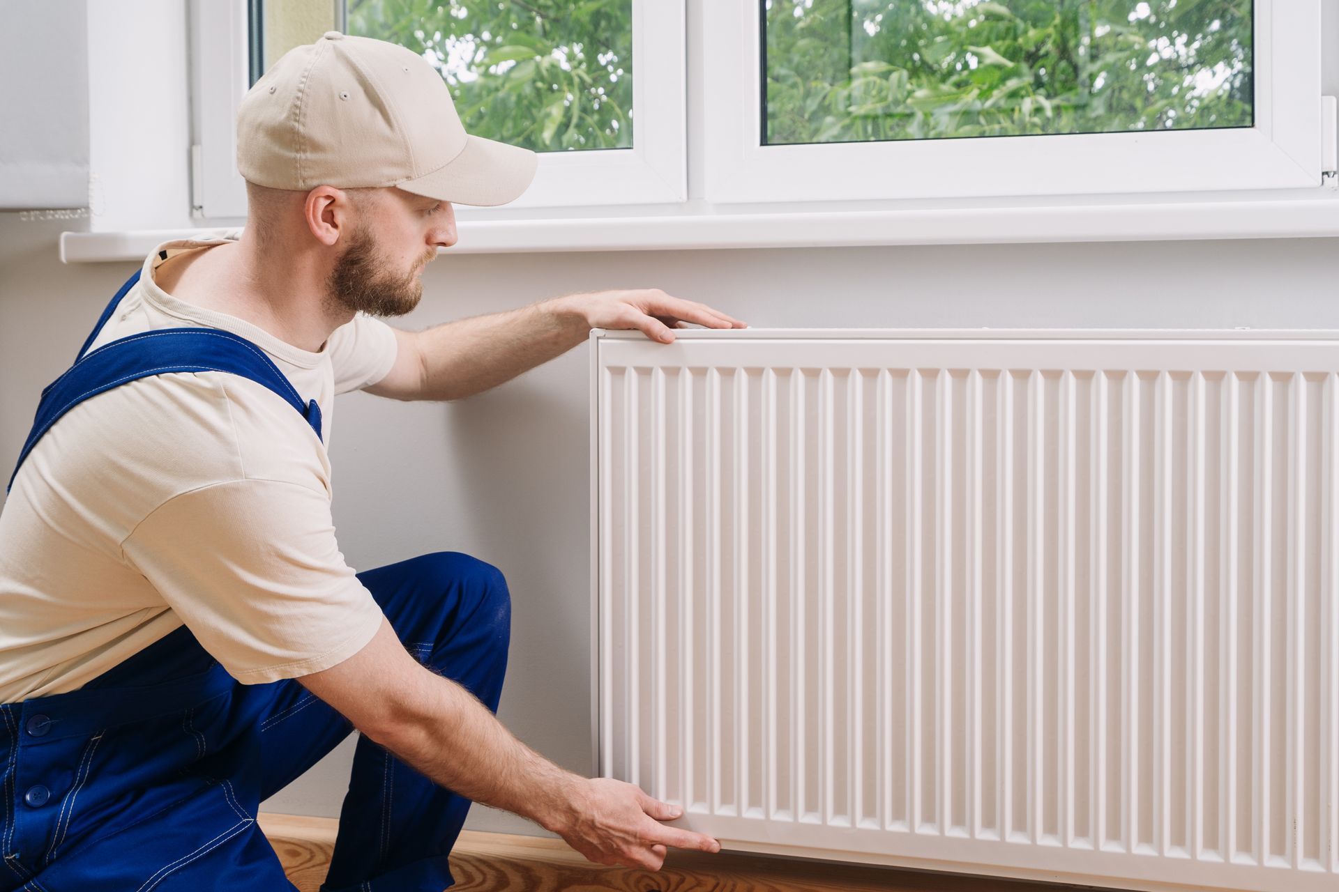 A man adjusting a wall-mounted radiator beneath a window during indoor heating system repair