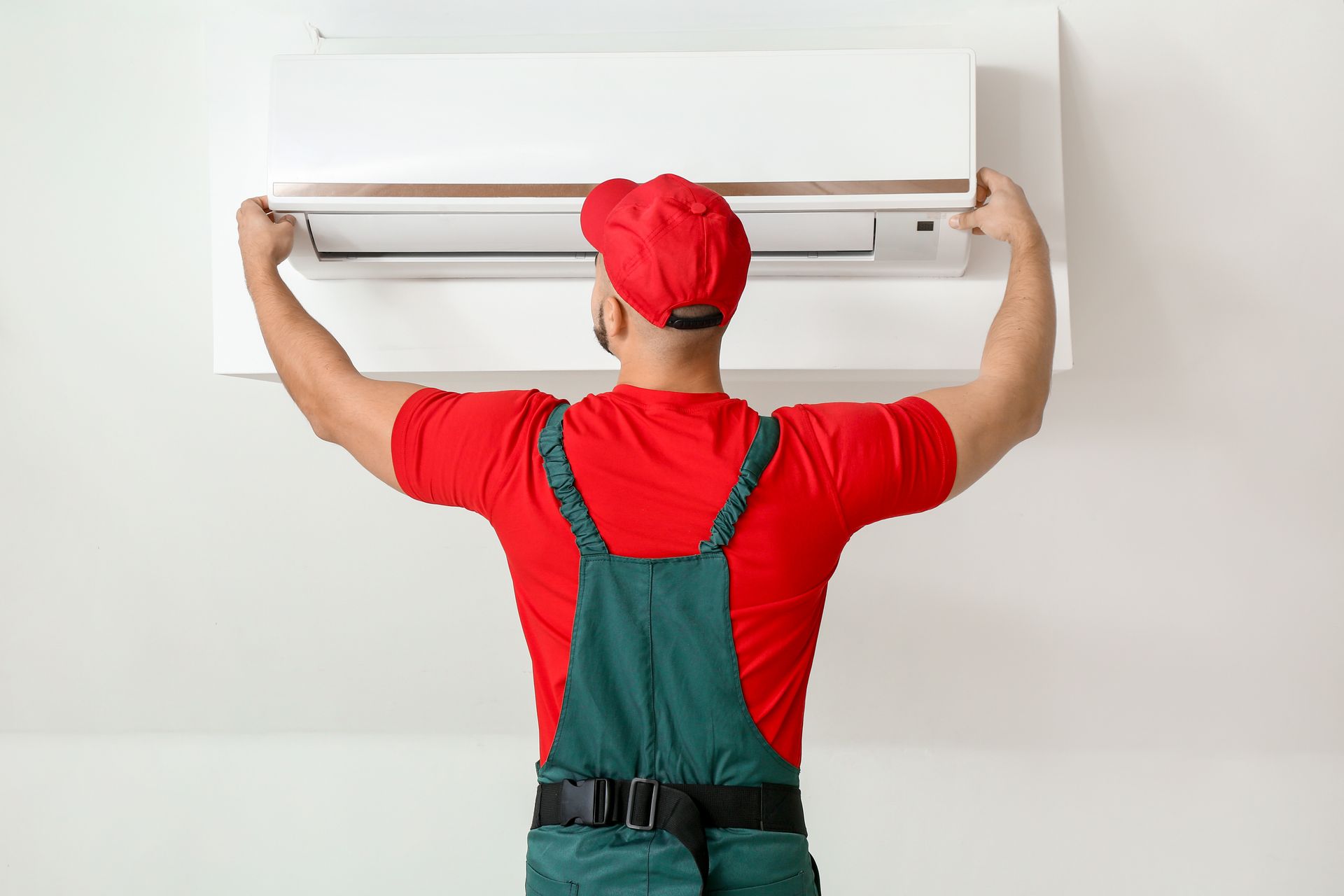 A technician in a red shirt and green overalls performs an air conditioning installation