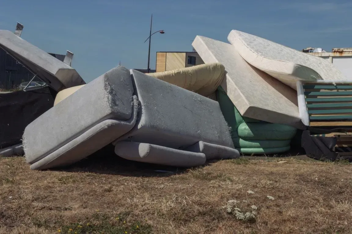 Pile of discarded mattresses and foam cushions outdoors on bare ground under a blue sky.