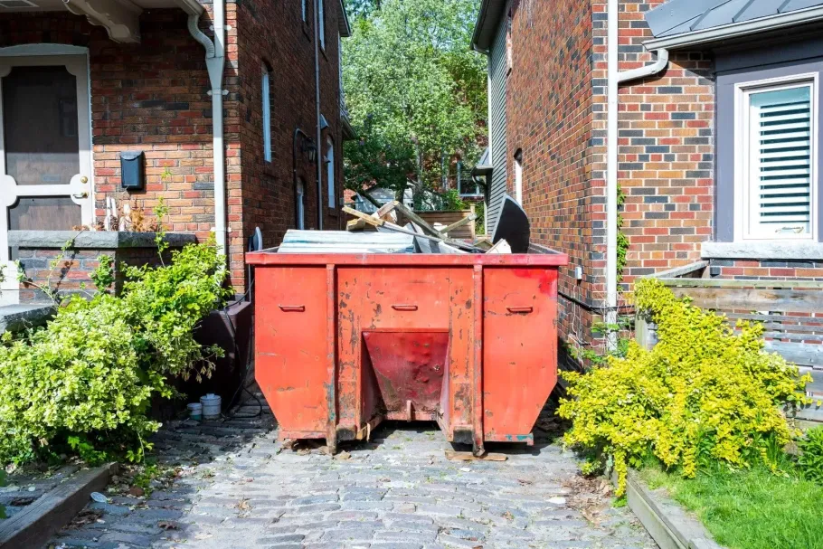 Large red dumpster between brick houses on a residential driveway, with bushes on both sides