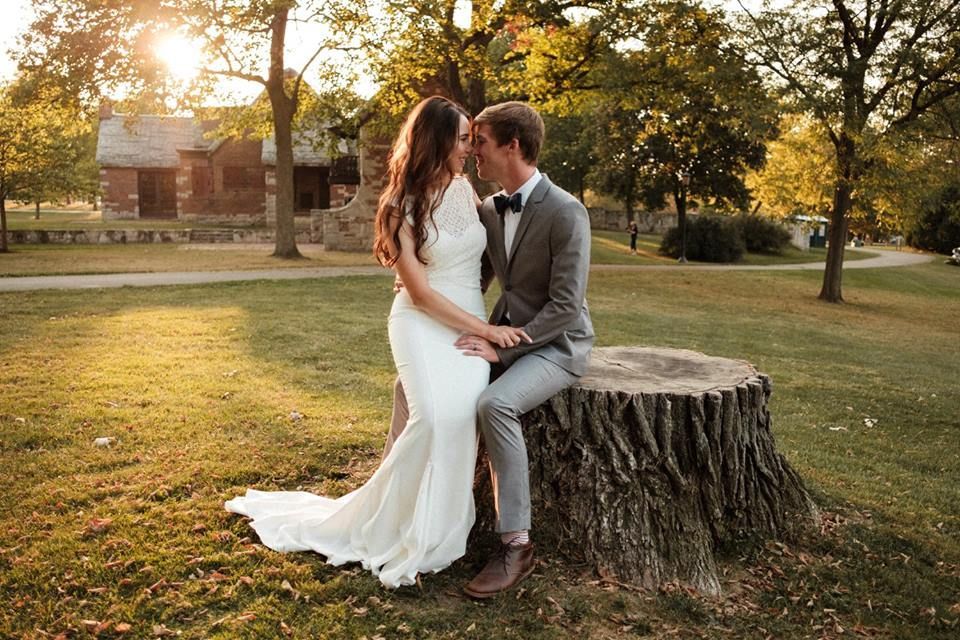 A bride and groom are sitting on a tree stump in a park.