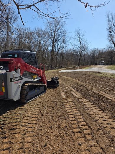 Red skid steer grading a dirt path on a sunny wooded hillside