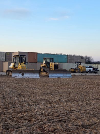 Three bulldozers on a muddy construction site beside stacked shipping containers under a cloudy sky