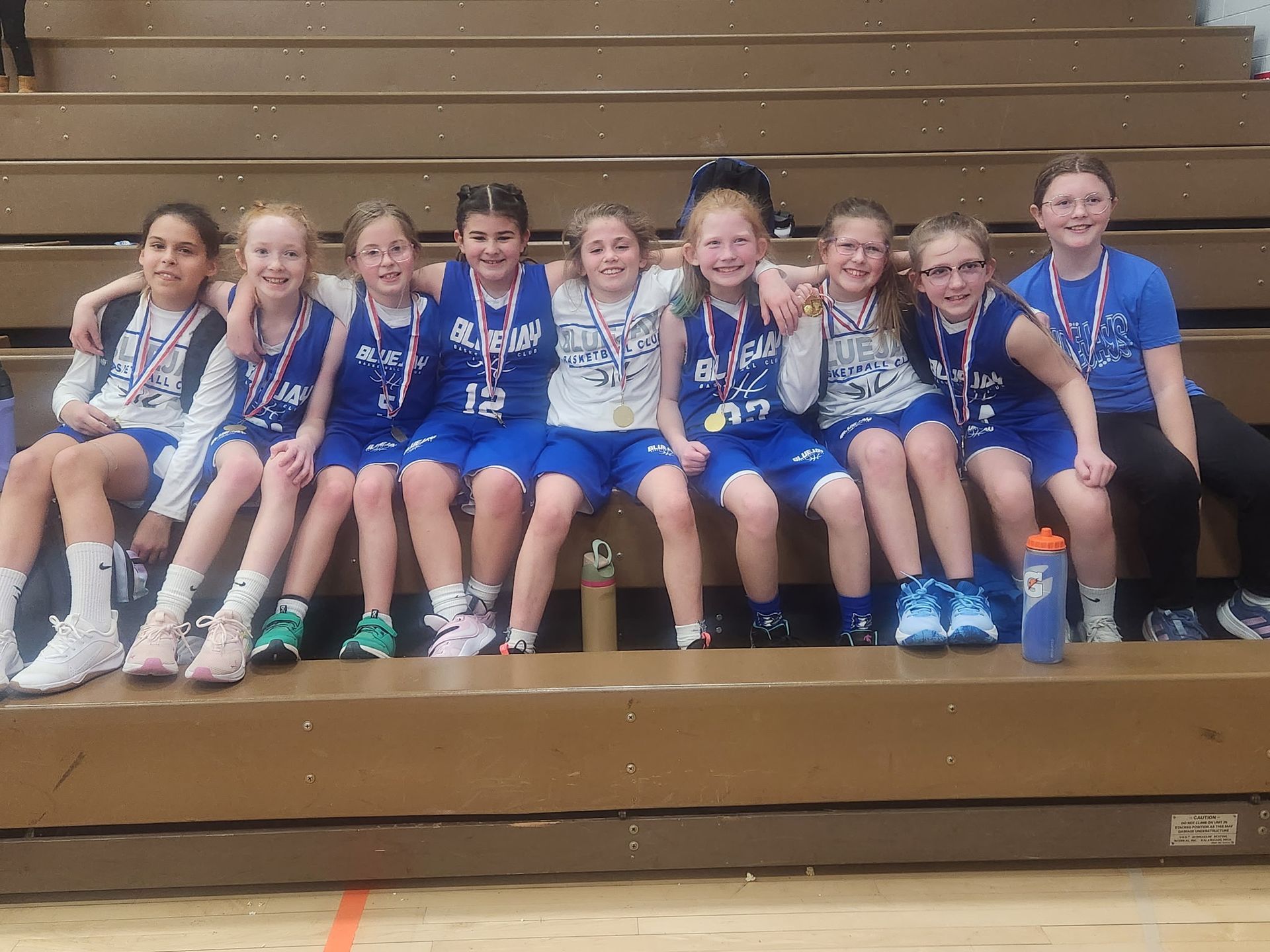 Youth sports team in blue uniforms sitting on bleachers, smiling with medals around their necks.