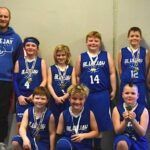 Youth basketball team posing in blue jerseys with medals, standing indoors against a plain wall
