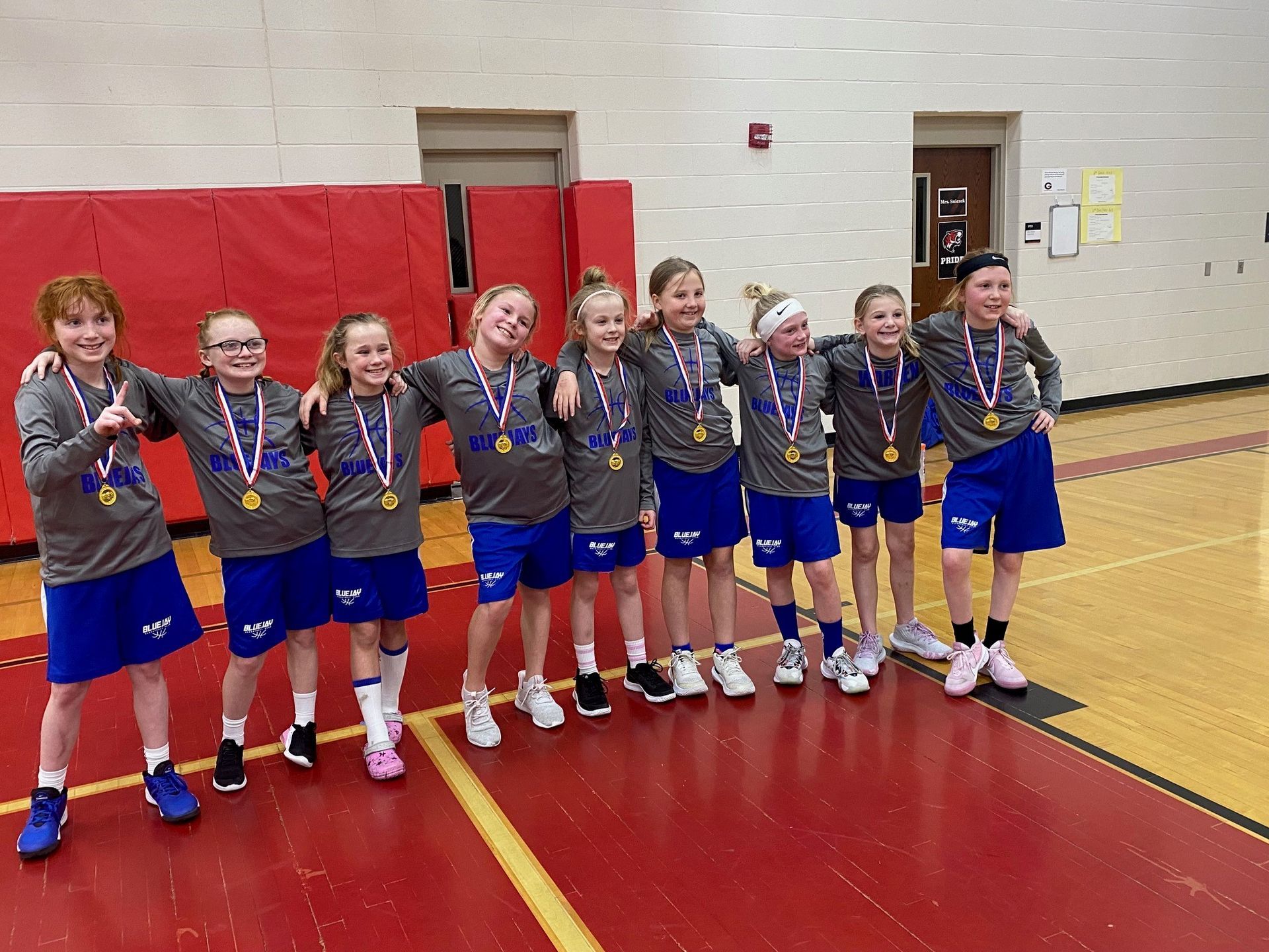Girls' basketball team posing in a gym, wearing medals and matching uniforms, smiling by red wall padding