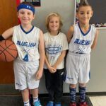 Three youth basketball players in white uniforms posing indoors, one holding a basketball.
