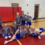 Youth soccer team posing on gym floor with medals, red mat in background.
