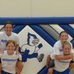 Cheerleaders posing in front of a blue-and-white mascot mural at a gym.
