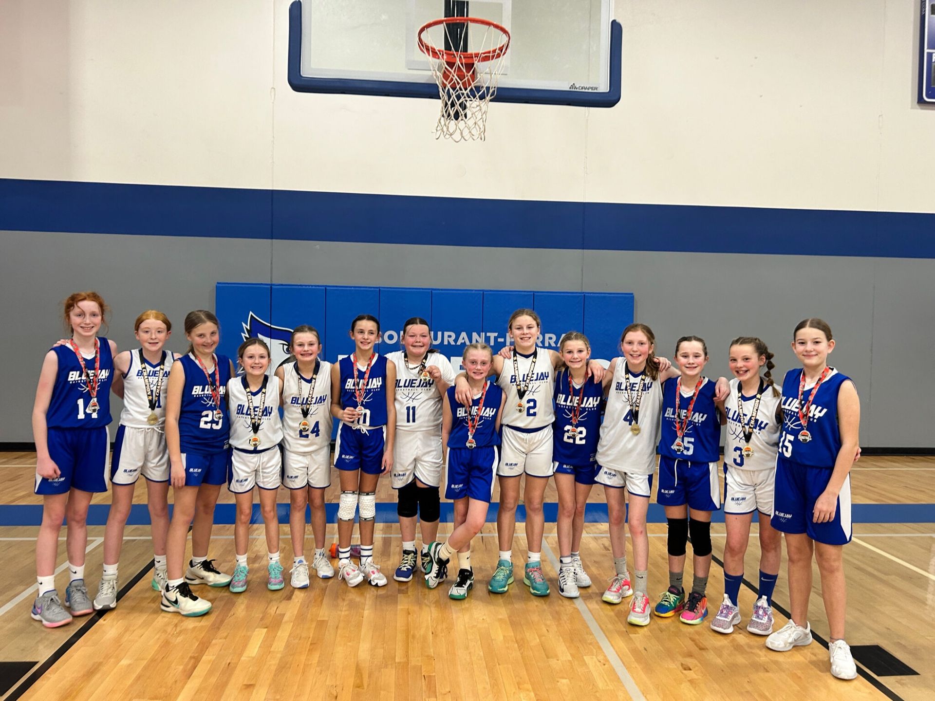 Youth basketball team posing on a gym court beneath the hoop, wearing blue and white uniforms.