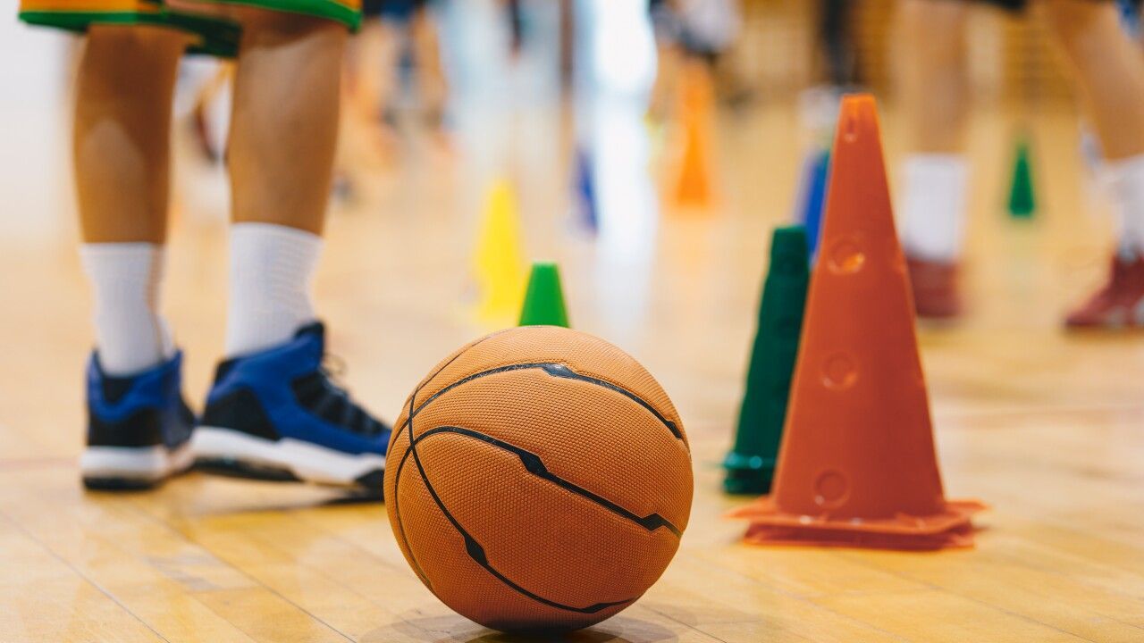Basketball on a gym floor beside orange cones, with a child's legs and sneakers in the background