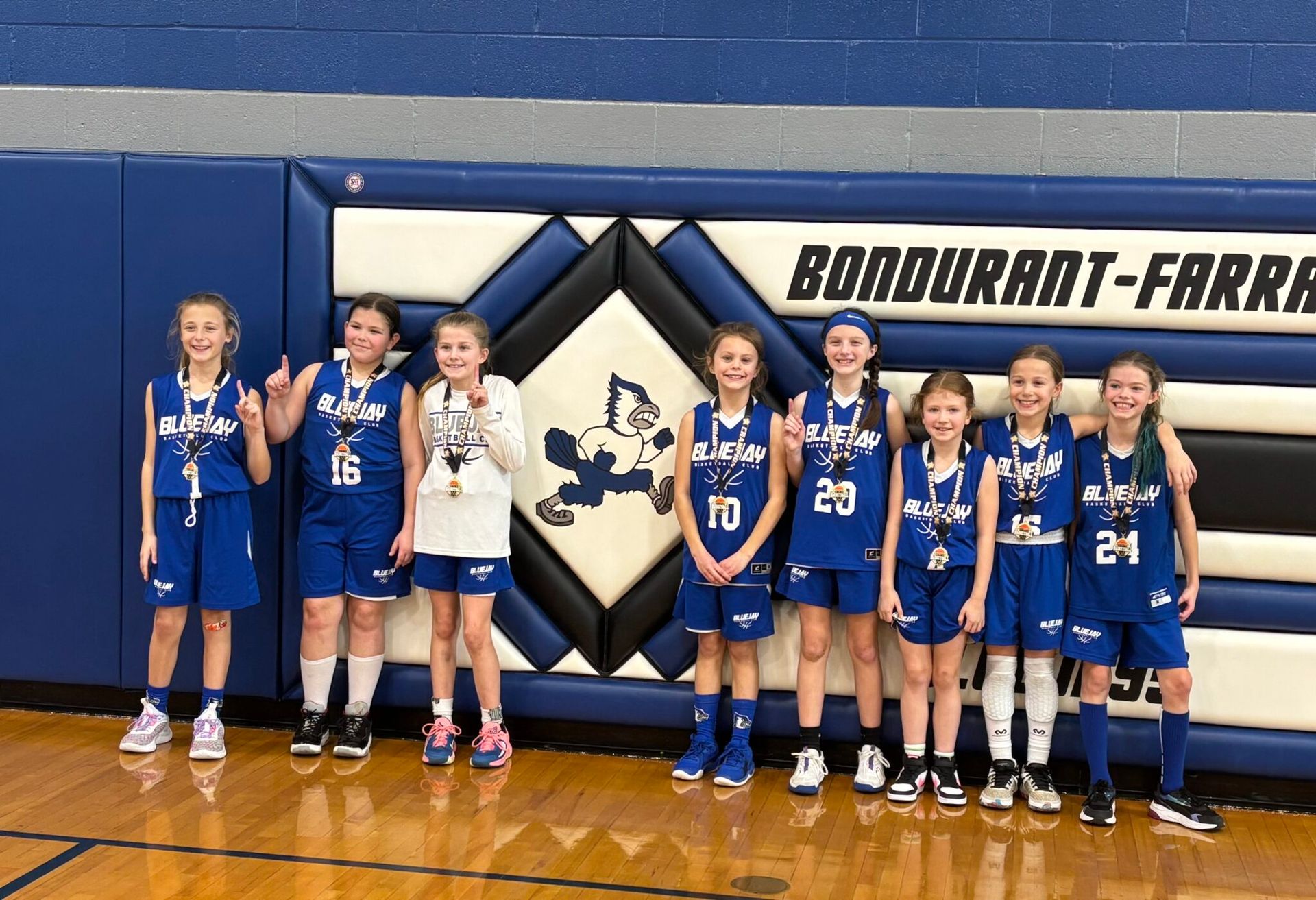 Youth sports team posing on a gym floor with medals and trophies, red wall pads in the background.