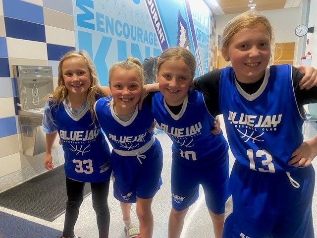 Four youth basketball players in blue jerseys pose smiling in a gym hallway