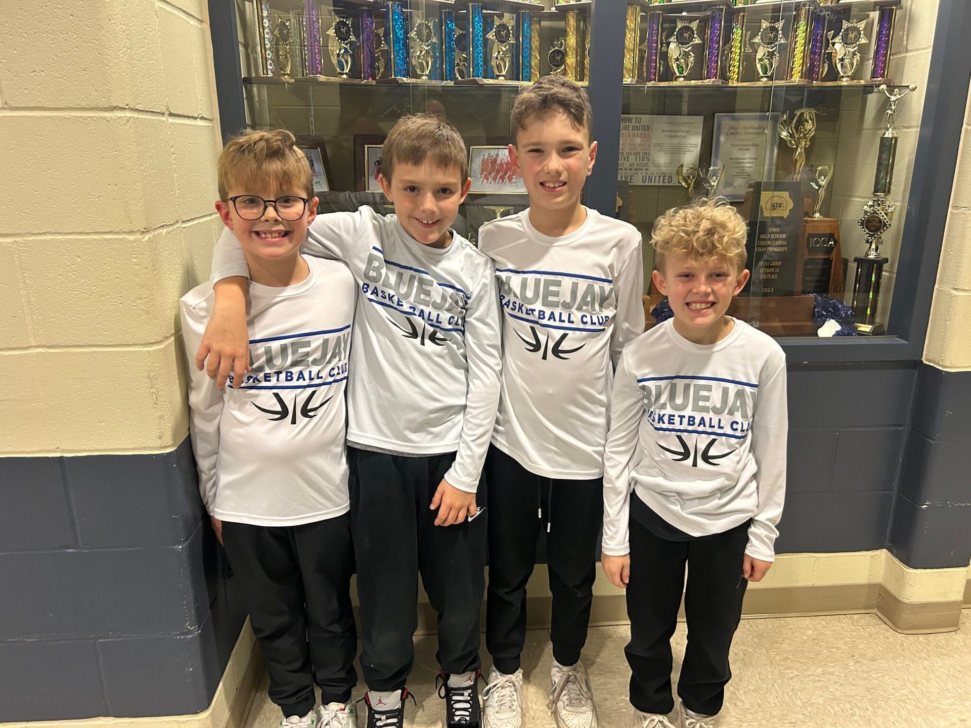 Four children in matching white shirts posing in front of a trophy case at school