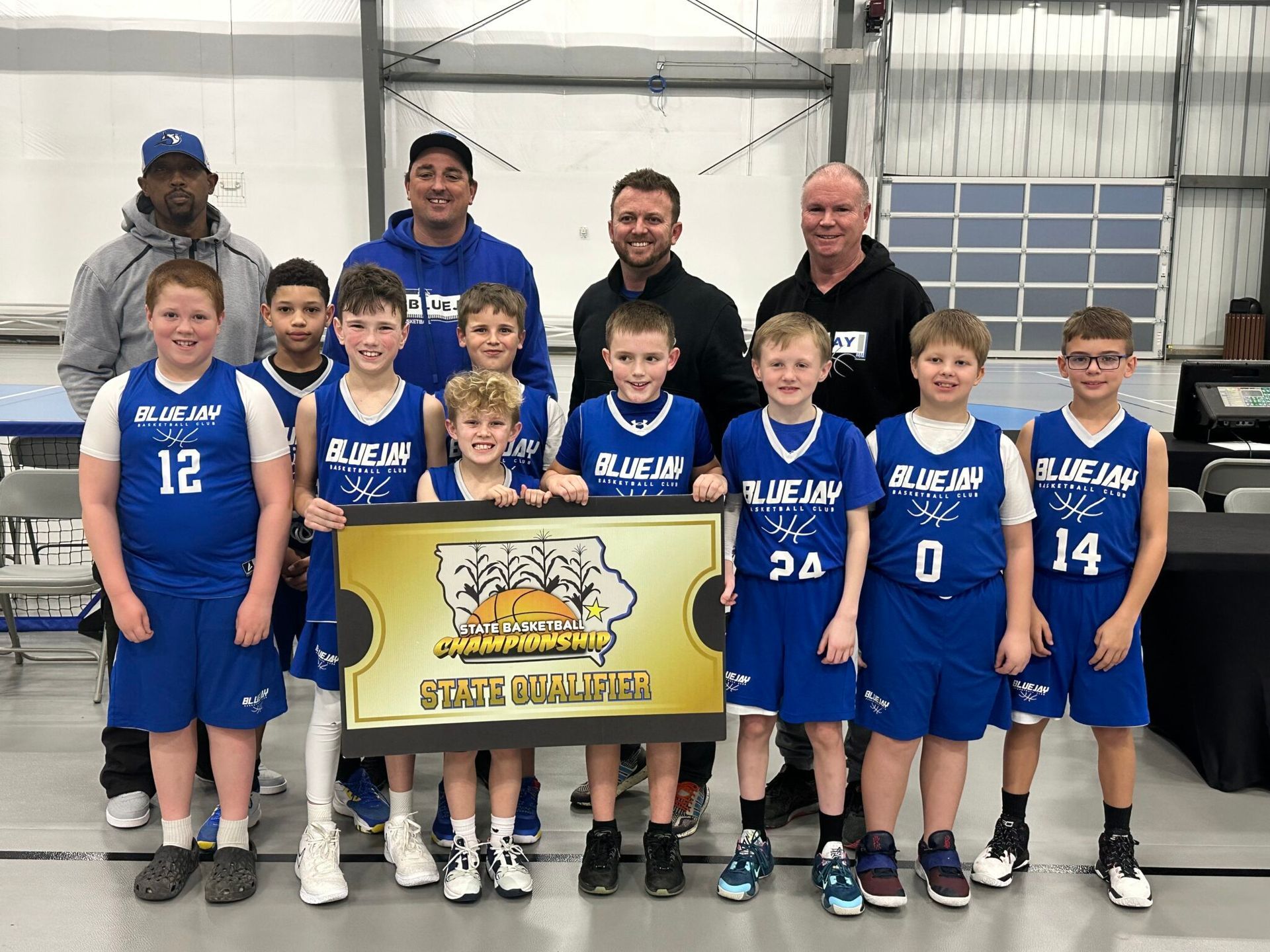 Youth basketball team in blue uniforms posing with coaches indoors, holding a championship banner.