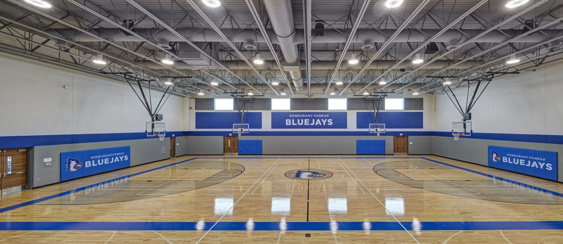 Indoor volleyball court with blue walls, wooden floor, and empty bleachers under bright lights