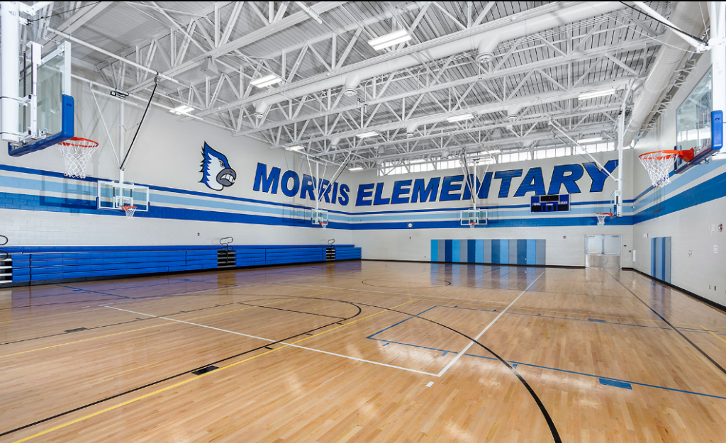 Empty elementary school gymnasium with polished wood floor and blue-and-white walls reading “MORRIS ELEMENTARY.”