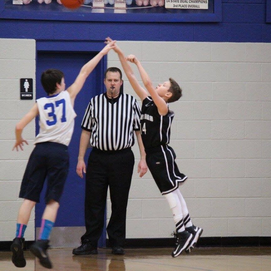 Two basketball players jump for a tip-off beside a referee on a gym court.