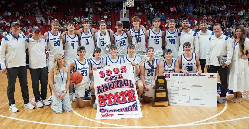 Basketball team posing with championship banner and trophy on the court after a state title win
